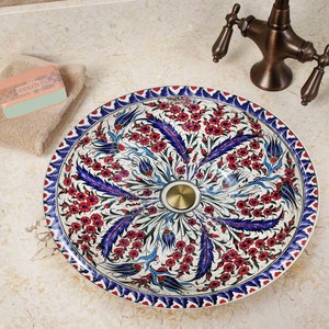 Puede incluir: Un lavabo de cerámica blanca con un patrón floral azul y rojo. El lavabo es redondo y tiene una tapa de drenaje decorativa de oro.