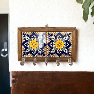 May include: A wooden key holder with a decorative tile featuring a blue, yellow and green floral pattern. The holder has five hooks for hanging keys.