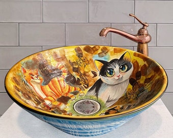May include: A yellow and blue ceramic bathroom sink with a cat and a bird design. The sink is on a white countertop with a copper faucet and a round mirror above it.