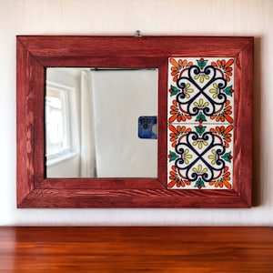 Puede incluir: Un espejo con marco de madera oscura con un diseño de azulejos decorativos en tonos de azul, amarillo, naranja y verde. El espejo está colgado en una pared blanca sobre una superficie de madera.