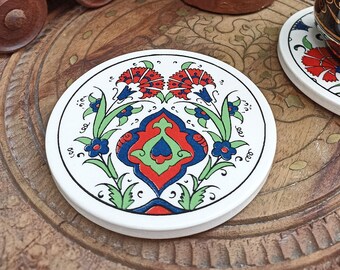 May include: Set of four white ceramic coasters with a colorful floral design. The coasters feature a red, green, blue, and white floral pattern with a geometric center. The coasters are displayed on a wooden surface with a brown ceramic lantern and a cup.