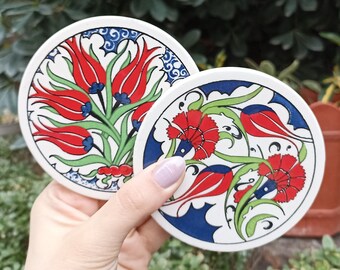 Puede incluir: Dos posavasos redondos de cerámica con fondo blanco y un diseño floral colorido. Los posavasos presentan flores rojas, verdes, azules y blancas con detalles intrincados.
