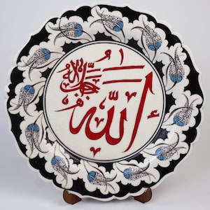 Puede incluir: Un plato de cerámica blanco con un diseño floral blanco y negro alrededor del borde. El centro del plato presenta una inscripción caligráfica roja de la palabra "Allah" en árabe.