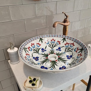 May include: A white ceramic bathroom sink with a floral design in blue, red, and green. The sink is on a white countertop with a black metal stand. There is a bar of soap on the countertop.