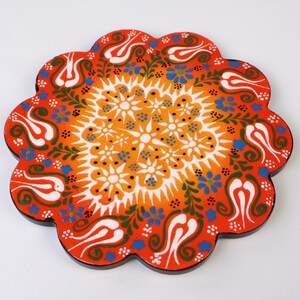 May include: A round, red, orange, white, green, and blue ceramic coaster with a floral design. The coaster has a scalloped edge.
