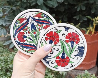Puede incluir: Dos posavasos redondos de cerámica con fondo blanco y un diseño floral colorido. El diseño presenta claveles rojos, hojas verdes y remolinos azules y blancos. Los posavasos son perfectos para proteger las superficies del calor y los derrames.