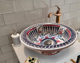 Puede incluir: Un lavabo de cerámica blanca con un diseño floral azul y rojo. El lavabo está sobre una encimera blanca con un soporte de metal negro. Hay un dispensador de jabón y una barra de jabón en la encimera.