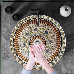 Puede incluir: Un lavabo de cerámica blanca con un patrón floral marrón y azul. El lavabo es redondo y tiene un grifo negro. El lavabo está colocado sobre una encimera gris.