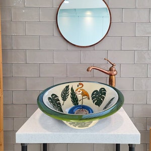 Puede incluir: Un lavabo de baño de cerámica blanca con un borde verde y un estampado tropical con un flamenco y hojas de palma. El lavabo está sobre una encimera blanca con un grifo de cobre.