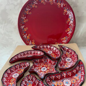 Może przedstawiać: Zestaw czerwonych ceramicznych naczyń do serwowania z białymi i pomarańczowymi wzorami kwiatowymi. Zestaw zawiera dużą okrągłą paterę i kilka mniejszych naczyń w kształcie półksiężyca i gwiazdy.