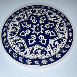 May include: A round ceramic coaster with a blue and white floral design. The coaster features a central starburst pattern surrounded by a border of stylized flowers and leaves.