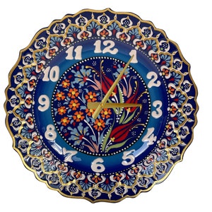 May include: Decorative ceramic wall clock with a blue floral design. The clock face features white numerals and gold-colored hands. The rim is scalloped and adorned with gold accents.