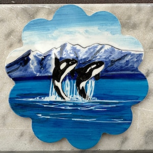Può includere: Un ornamento in legno dipinto a mano che raffigura due orche che saltano fuori dall'acqua davanti a una catena montuosa innevata. Lo sfondo è un cielo blu con nuvole bianche.