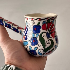 May include: A white ceramic coffee pot with a floral design in shades of blue, red, and green. The pot has a curved handle and a rounded body.