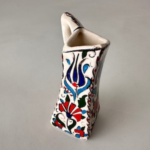 Puede incluir: Un jarrón de cerámica blanca con forma triangular y asa. El jarrón está decorado con un patrón floral colorido en tonos azules, rojos, verdes y blancos.