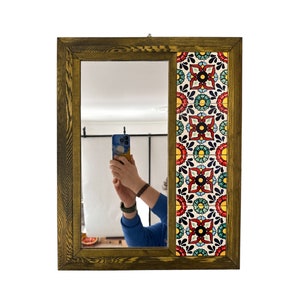 Puede incluir: Un espejo con marco de madera y un diseño de azulejos coloridos en el lado derecho. El espejo refleja a una persona tomando una selfie con un teléfono inteligente.