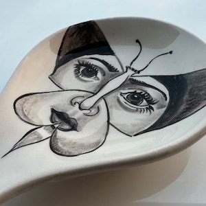 May include: A white ceramic spoon rest with a black and white illustration of a butterfly with two faces. The butterfly has a long, thin nose and a pair of large, black eyes.