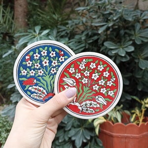 May include: Two round ceramic coasters with a white background and floral designs. One coaster features blue and green floral designs with red accents. The other coaster features red and green floral designs with white accents.