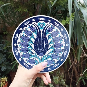 May include: A round ceramic plate with a blue and white floral design. The design features a central tulip with smaller tulips surrounding it. The plate has a white background with a blue border.