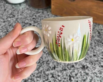 May include: White ceramic mug with a hand-painted design of white daisies and green grass. The daisies have yellow centers.