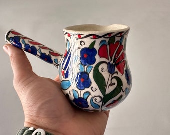 May include: A white ceramic coffee pot with a floral design in shades of blue, red, and green. The pot has a curved handle and a rounded body.