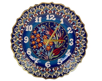 May include: Decorative ceramic wall clock with a blue floral design. The clock face features white numerals and gold-colored hands. The rim is scalloped and adorned with gold accents.