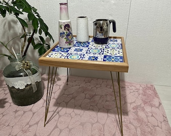 May include: A small square table with a tiled top featuring blue, white, and yellow geometric patterns. The table has four gold metal hairpin legs and sits on a pink rug. There are three decorative items on the table: a blue and white patterned vase, a white vase, and a silver teapot.