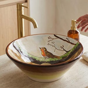 Peut inclure: Une vasque ronde en céramique ornée d'un rouge-gorge peint à la main perché sur une branche sur fond clair. L'extérieur du lavabo présente un dégradé allant d'un bord brun foncé à une base jaune clair. Un robinet doré brossé est installé.