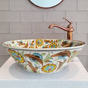 May include: A decorative ceramic sink with a floral pattern in yellow, blue, and brown sits atop a white countertop. A copper faucet and a round mirror with a copper frame are mounted on the gray tiled wall.