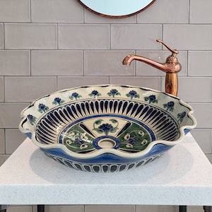 Op de afbeelding: Een witte keramische wastafel met blauwe en gouden bloemmotieven. De wastafel staat op een wit aanrecht met een koperen kraan en een ronde spiegel erboven.