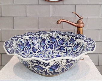 May include: A white ceramic sink with a blue and white floral pattern. The sink has a wavy edge and is sitting on a white countertop. A copper faucet is attached to the sink. A round mirror hangs on the wall behind the sink.