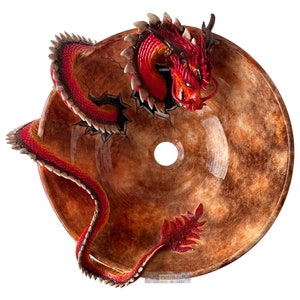 May include: A decorative brown glass sink featuring a red and black dragon sculpture. The dragon is depicted emerging from the sink's surface. The sink is circular with a central drain hole.