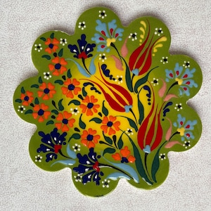 May include: A green ceramic coaster with a floral design. The design features red, yellow, orange, blue, and white flowers on a green background.