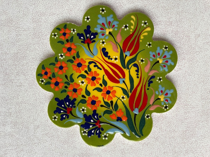 May include: A green ceramic coaster with a floral design. The design features red, yellow, orange, blue, and white flowers on a green background.