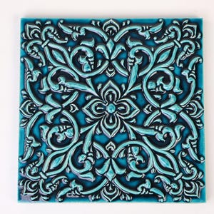 Puede incluir: Un azulejo de cerámica azul turquesa con un intrincado diseño floral en relieve. El diseño es de color azul oscuro y presenta un patrón repetitivo de hojas y flores estilizadas.