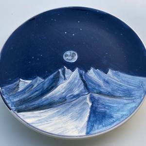 Op de afbeelding: Een blauw keramiek bord met een handgeschilderd ontwerp van een bergketen onder een sterrenhemel met volle maan.