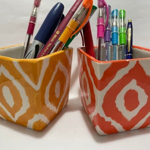 May include: Two ceramic pencil holders with geometric patterns. The holder on the left is orange with white geometric shapes. The holder on the right is coral with white geometric shapes. Both holders are filled with pens and pencils.