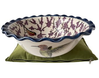 May include: A white ceramic bowl with a blue rim and a floral design featuring pink blossoms and two birds. The bowl is sitting on a green cushion with a zipper.