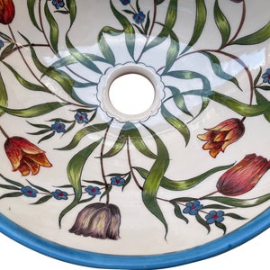 May include: A white ceramic sink with a blue rim and a floral design. The sink has a drain hole in the centre and is decorated with green leaves, orange and red tulips, and blue flowers.