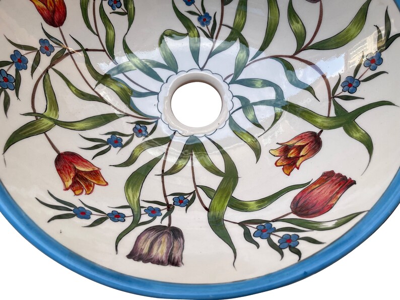 May include: A white ceramic sink with a blue rim and a floral design. The sink has a drain hole in the centre and is decorated with green leaves, orange and red tulips, and blue flowers.
