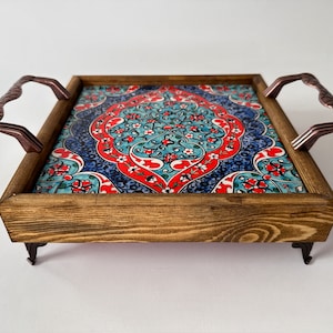 May include: A wooden serving tray with ornate tile inlay in shades of blue, red, and white. The tray has two metal handles and four small feet.