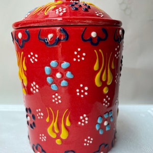 Op de afbeelding: Rode keramische pot met een deksel, versierd met een kleurrijk bloemenpatroon. De pot heeft een witte achtergrond met gele, blauwe en zwarte accenten.