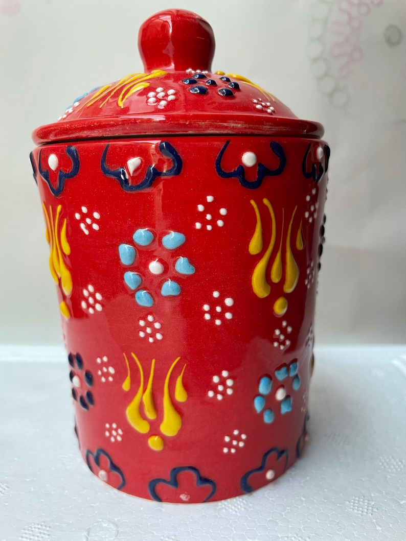 Op de afbeelding: Rode keramische pot met een deksel, versierd met een kleurrijk bloemenpatroon. De pot heeft een witte achtergrond met gele, blauwe en zwarte accenten.