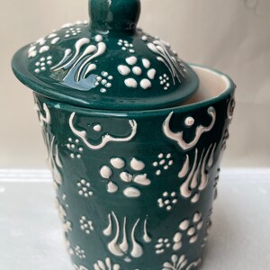 Op de afbeelding: Een groene keramische pot met een deksel, beide versierd met witte bloemmotieven. De pot heeft een rond lichaam en een brede opening.