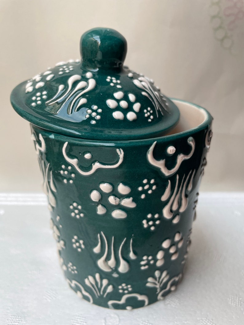 Op de afbeelding: Een groene keramische pot met een deksel, beide versierd met witte bloemmotieven. De pot heeft een rond lichaam en een brede opening.