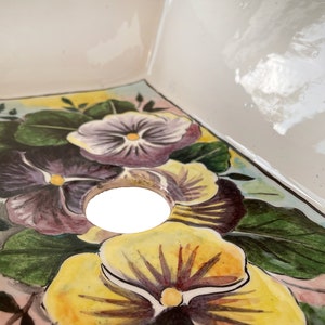 May include: A white ceramic sink with a square shape and a drain hole. The sink is decorated with hand-painted purple, yellow, and green pansies.