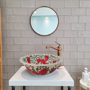 May include: A white ceramic bathroom sink with a floral design featuring red roses and green leaves. The sink is on a white countertop with a copper faucet and a round mirror above it.