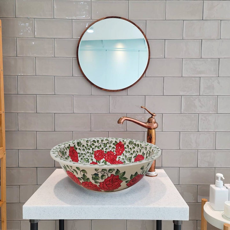 May include: A white ceramic bathroom sink with a floral design featuring red roses and green leaves. The sink is on a white countertop with a copper faucet and a round mirror above it.