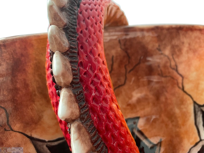 May include: Close-up of a ceramic mug with a detailed dragon design. The mug features a vibrant red interior with textured scales, a row of white teeth, and a brown exterior with a cracked pattern.