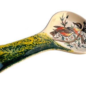 May include: A ceramic spoon rest with a green and yellow glaze. The top of the rest features a painted design of two birds perched on a branch with white flowers.